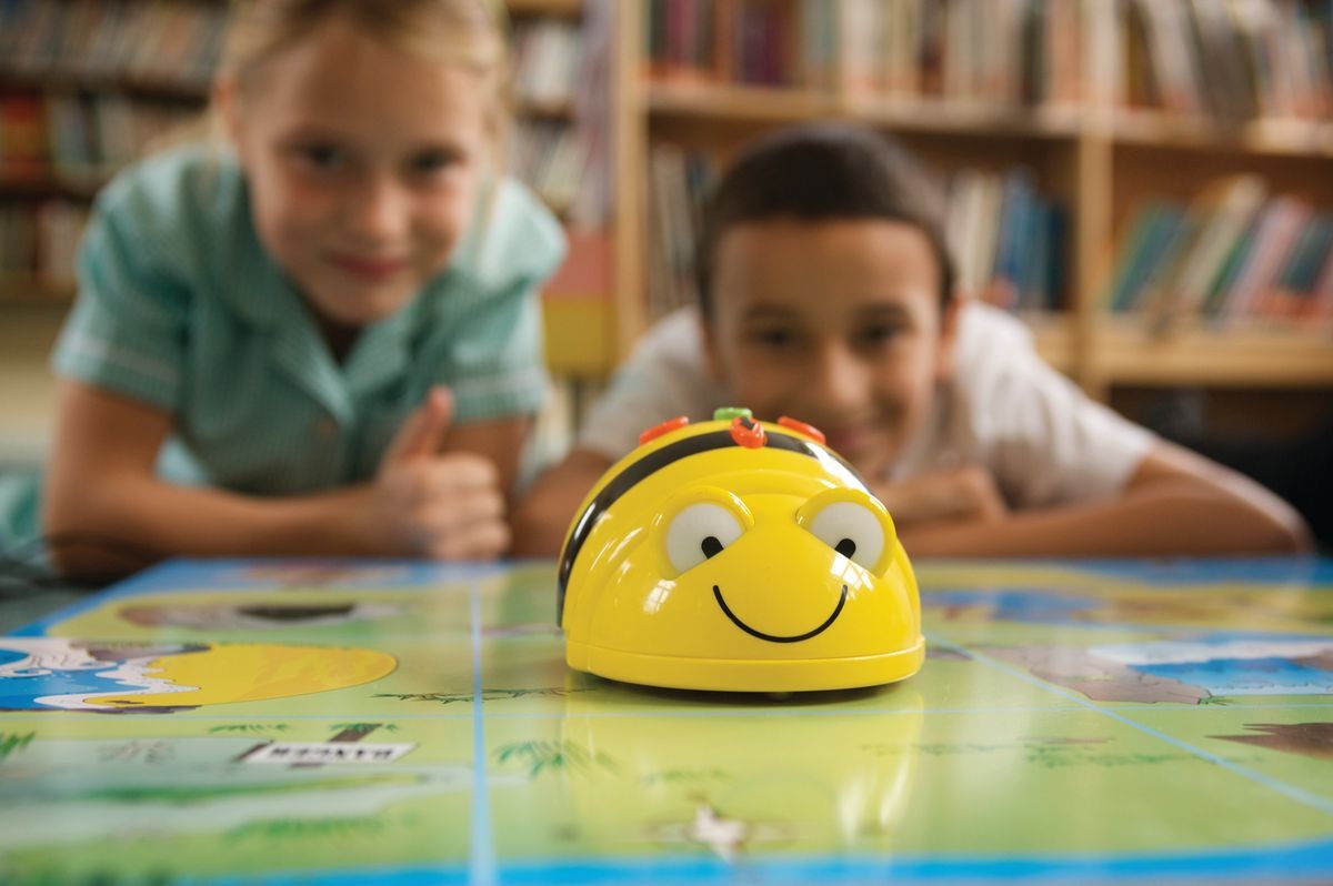 A photo of bookshelf with two children in front of it looking at a bee shaped robot on a table.