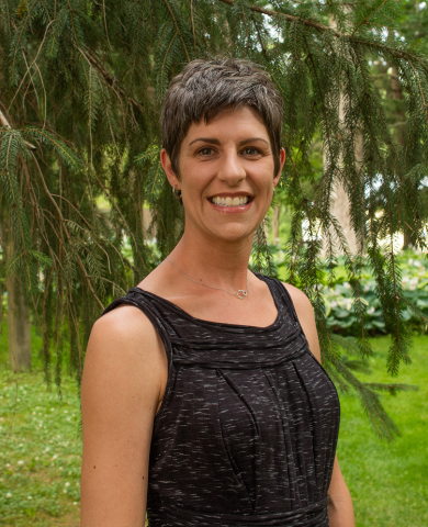 Photo of the presenter, Jessi Bushman standing outside in front of coniferous branches. Jessi is a tall white woman with short dark hair.