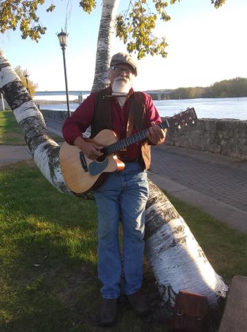 Picture of the musician Denny Garcia standing in front of a bent white tree and a brick trail overlooking the Mississippi River in the background. Denny is holding an acoustic guitar and wearing a harmonica around his neck. 