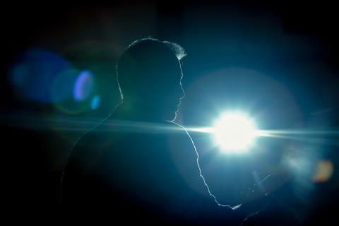 Photo of a flare of light in a dark stage illuminating an indistinct man facing away from the viewer and toward the audience. It appears he's giving a speech of some kind. 