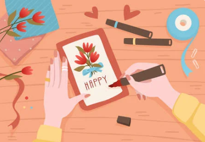 An image of a pair of hands creating a card that says "Happy" with flowers on top of it with a calligraphy pen. There are also several cardmaking supplies on the  table like washi tape, flowers, and paper.