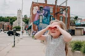 A young girl standing on a city sidewalk with her hands around her eyes like binoculars.