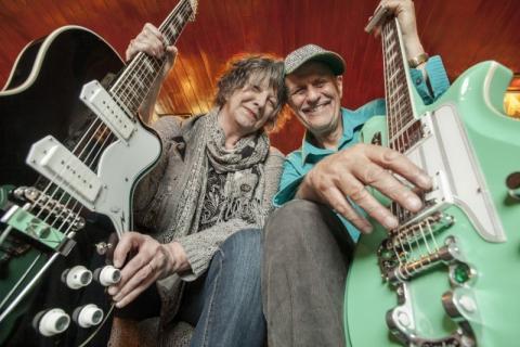 Colorful low-view photograph of the husband and wife duo Joe and Vicki Price holding their electric guitars.