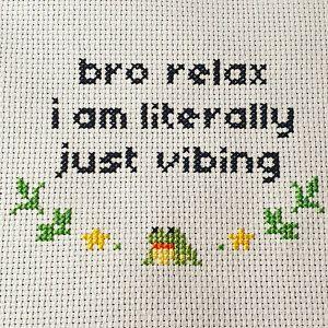 Black cross stitched letters that read "bro relax i am literally just vibing" with a small frog and leaf decorations underneath