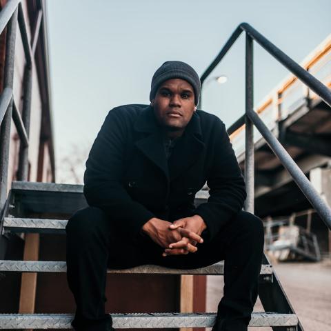 Photo of the artist Soultru sitting outside on the stairs. Soultru is a young african american male. In the picture he's wearing a jacket and hat.