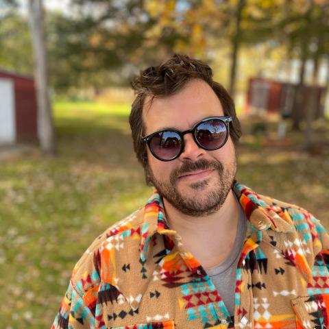 Photograph of singer/songwriter Joel Sires. Sires is standing outside on a sunny day wearing sunglasses.