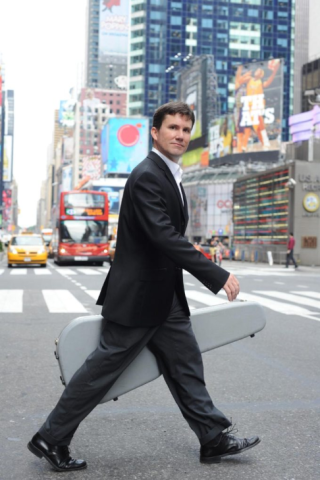 Photo of classical guitarist Peter Fletcher wearing a black suit and crossing a busy city street with his hardshell guitar case in hand. 