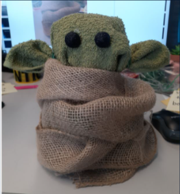 A photograph of a Baby Yoda plush made from a green towel and burlap ribbon set on a table.