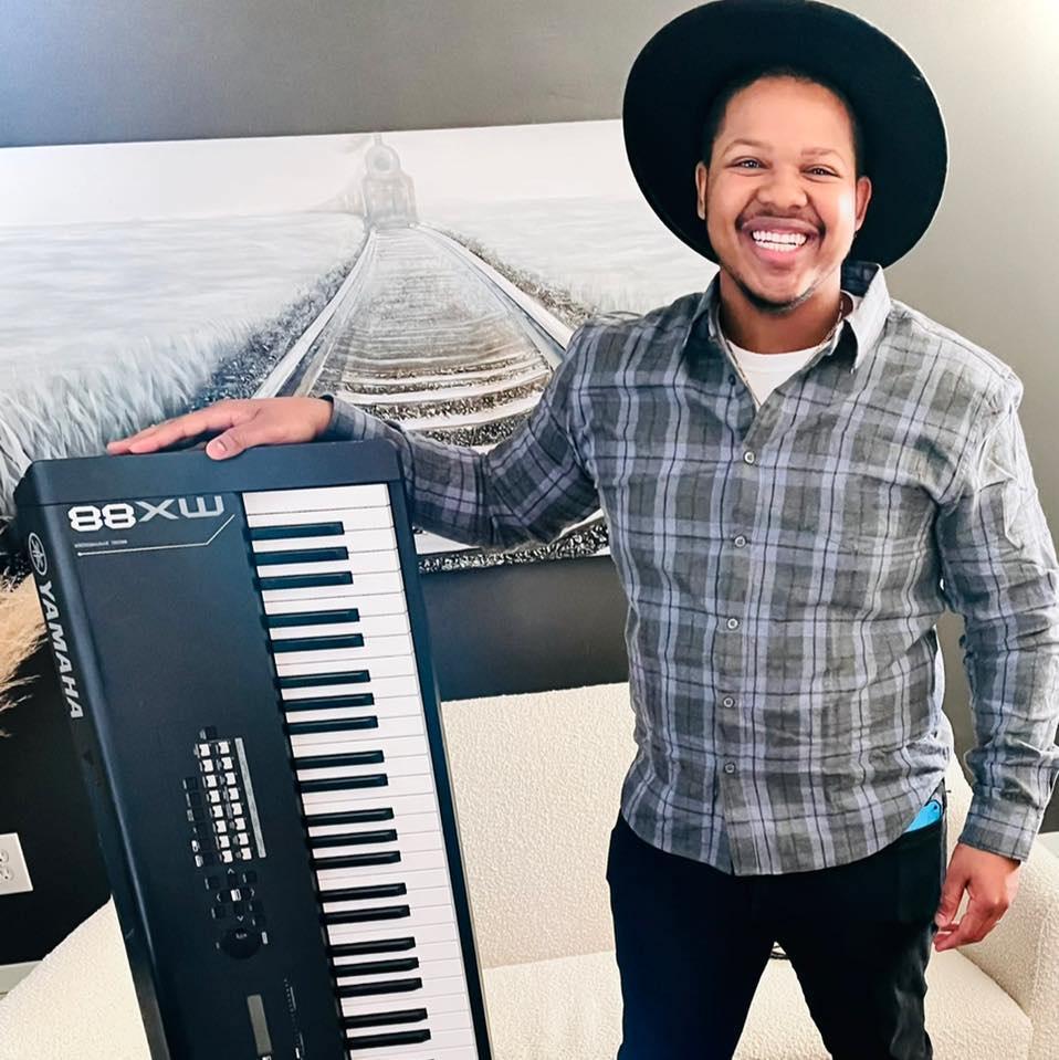 The musician, Brion Bowman smiling and standing with his electric keyboard in front of a painting of train tracks.