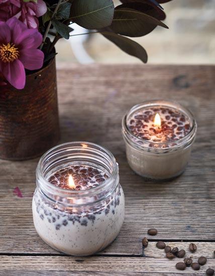 A photograph of two glass jars with wax, coffee beans, coffee grounds, and wicks, set on a table. 