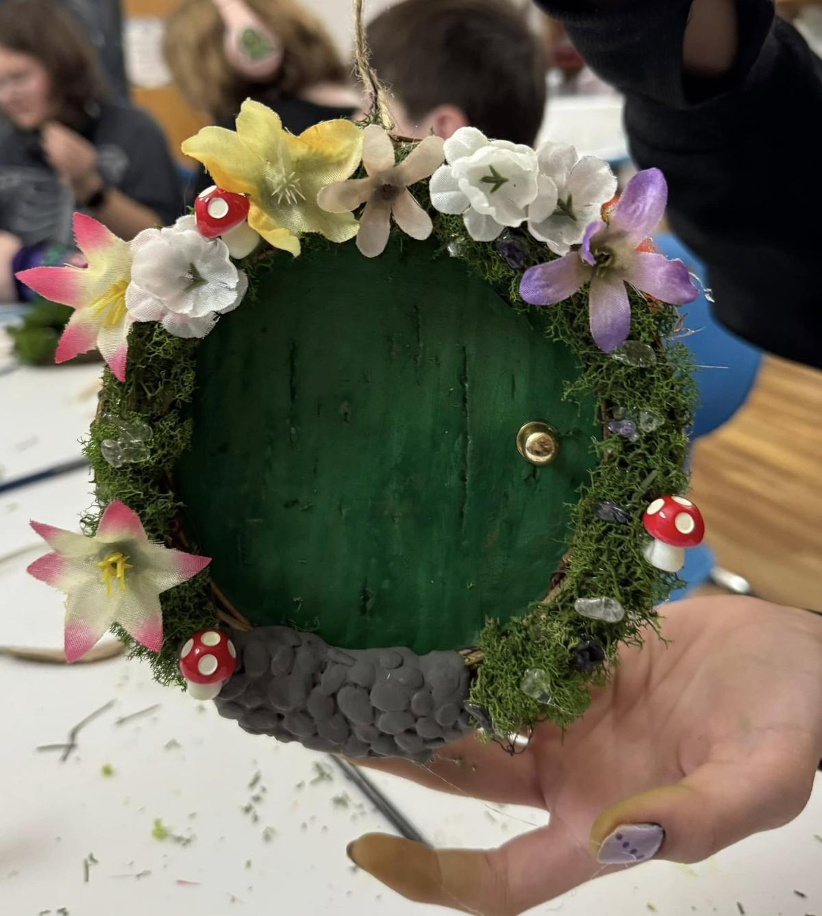 Photograph of a green hobbit door wreath, with flowers and mushrooms surrounding it. 